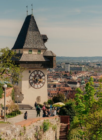 Grazer Uhrturm auf dem Schlossberg: Blick auf den Grazer Uhrturm mit Spaziergänger*innen auf dem Weg davor und Panoramablick über die Altstadt von Graz im Hintergrund.