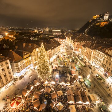 Blick über den festlich beleuchteten Christkindlmarkt in der Grazer Innenstadt bei Nacht. Zahlreiche Marktstände und ein großer, geschmückter Weihnachtsbaum prägen den Hauptplatz, im Hintergrund ist der hell erleuchtete Schlossberg mit Uhrturm zu sehen.