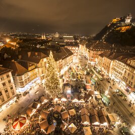 Blick über den festlich beleuchteten Christkindlmarkt in der Grazer Innenstadt bei Nacht. Zahlreiche Marktstände und ein großer, geschmückter Weihnachtsbaum prägen den Hauptplatz, im Hintergrund ist der hell erleuchtete Schlossberg mit Uhrturm zu sehen.