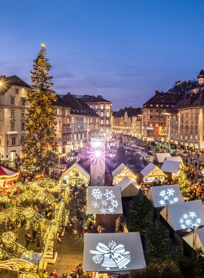 „Stimmungsvoll beleuchteter Adventmarkt in der Grazer Altstadt bei Einbruch der Dunkelheit, mit festlich geschmückten Ständen, einem großen Weihnachtsbaum und Blick auf den Uhrturm am Schlossberg.