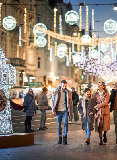 Four people walk along the festively lit Herrengasse with illuminated clocktower sculpture