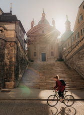 Woman cycling past the Mausoleum steps in Graz, with sunlight shining behind the dome