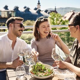 Drei Gäste genießen Speisen und Wein auf einer Terrasse mit Blick auf das Kunsthaus Graz.