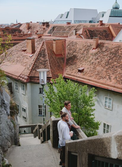 Two people standing on the Schlossberg Steps overlooking the rooftops of Graz's historic old town.