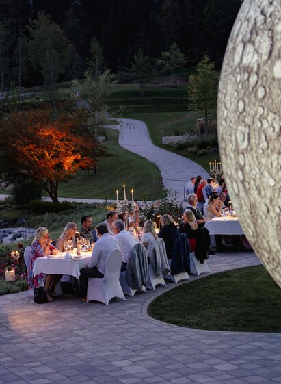 Guests enjoying dinner at the Vollmund culinary series at long tables in a park, lit by candles and light installations