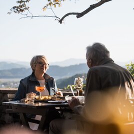 Zwei Personen genießen ein Essen auf einer Terrasse mit Panoramablick über die hügelige Landschaft der Südsteiermark im warmen Abendlicht.