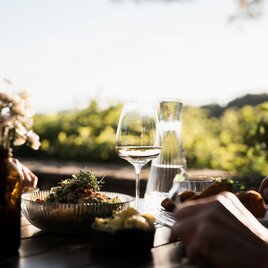 Gedeckter Tisch im Freien mit Weißwein, Aperitif und regionalen Speisen. Im Hintergrund sorgen Weinberge und warmes Abendlicht für eine stimmungsvolle Atmosphäre.