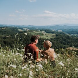 Paar sitzt inmitten einer Blumenwiese und blickt gemeinsam über die sanfte Hügellandschaft der Südsteiermark.