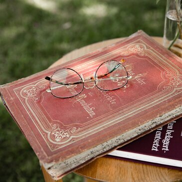 Buch mit rotem historischen Einband auf der eine runde goldene Lesebrille liegt.