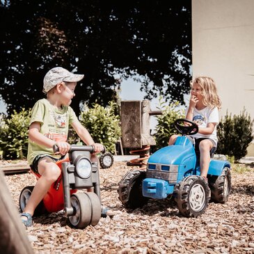 Zwei Kinder spielen auf einem Spielplatz mit einem roten Spielzeugmotorrad und einem blauen Trettraktor.