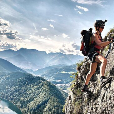 Frau klettert von der Seite auf Felsen, hinter ihr eine atemberaubende Aussicht auf Berge mit leicht wolkigem Himmel.