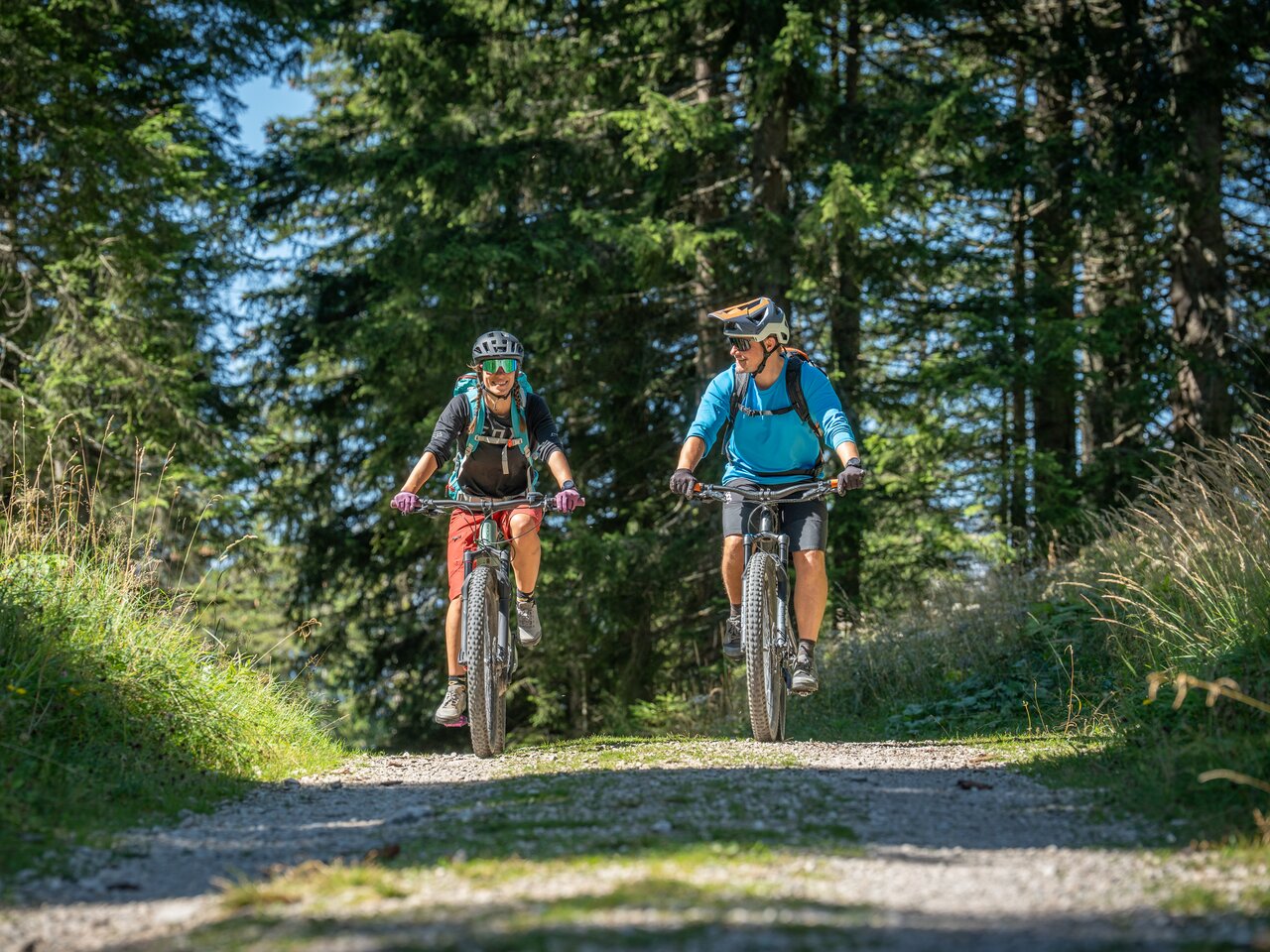 Zwei Radfahrer fahren der Kamera lachend entgegen auf einem Forstweg in einem Wald.
