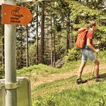 Im Vordergrund hängt ein Schild mit der Richtungsanweisung worauf ,,Styrian Iron Trial" steht, in diese Richtung gehen zwei Wanderer