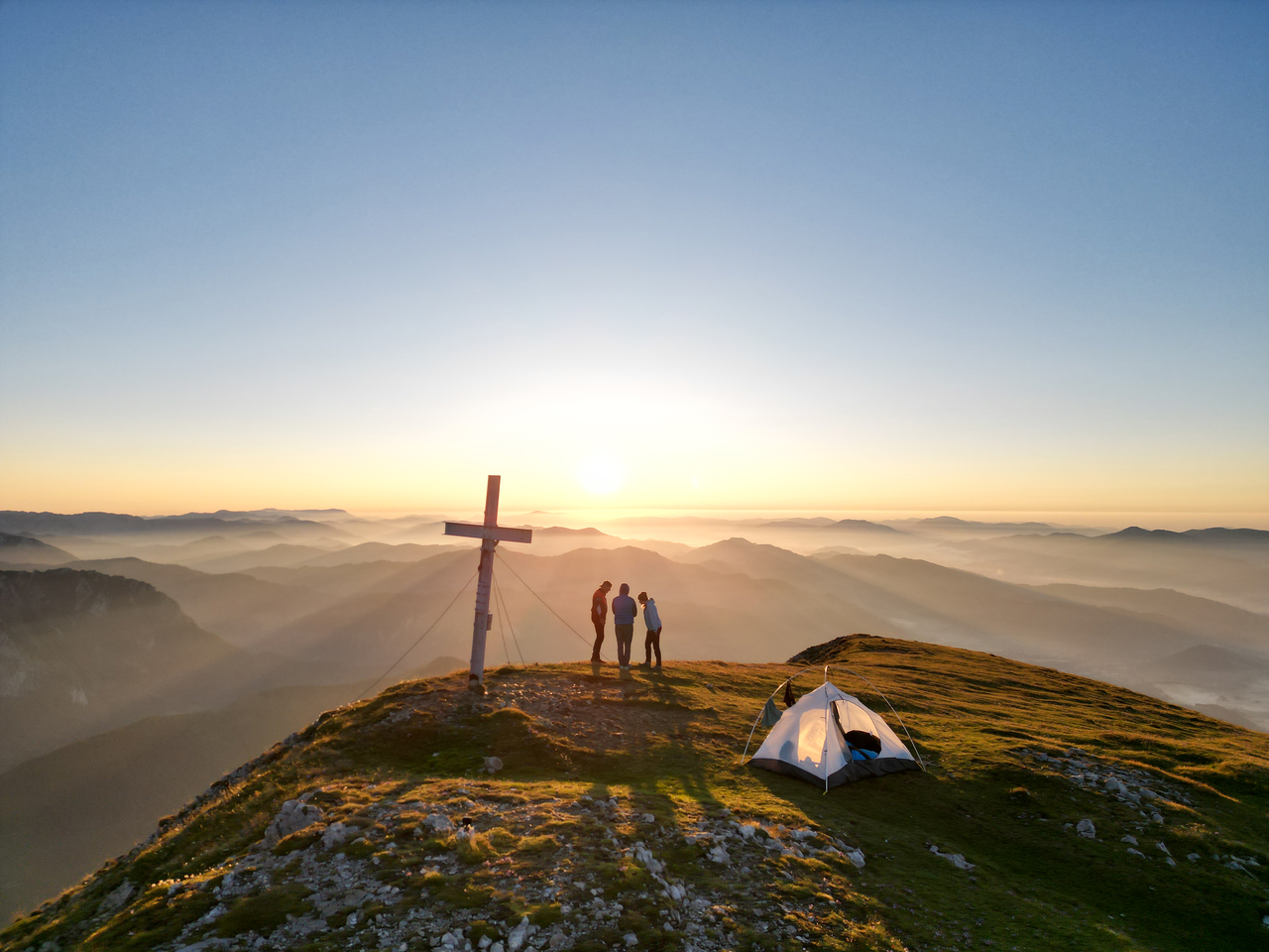 Drei Personen von hinten stehen neben dem Gipfelkreuz am Berg in einer Wiese neben einem Zelt und sie schauen dem Sonnenaufgang entgegen.