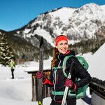Eine Frau mit Langlauf-Skiern in der Hand die vor winterlicher Kulisse an der Kamera vorbei lächelt.