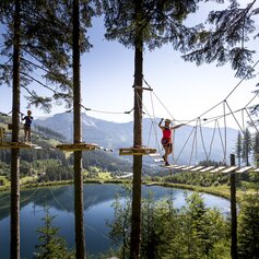 Zwei Personen klettern auf dem Adventure Park vor einem See in den Bäumen