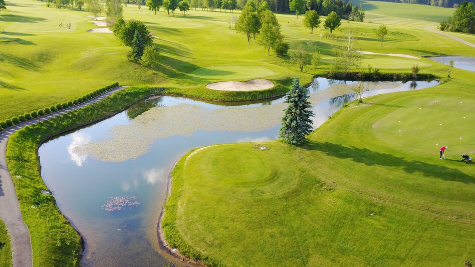 Golfplatz von oben mit Teich und dahinter vereinzelte Bäume am Wiesenrasen und dahinter sind Berge mit Wäldern.