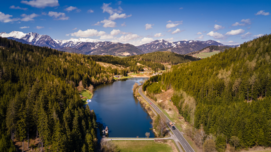 Trabochersee von oben mit umliegender Straße und dahinter eine Aussicht auf eine atemberaubende Bergkulisse