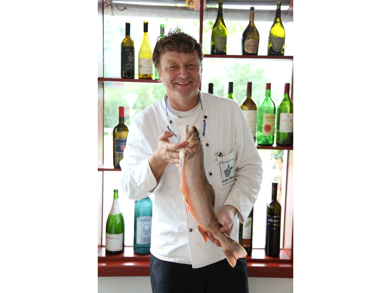 Klaus Lobnik in Kochgewand vor einer Wand mit Weinflaschen mit einem Fisch in der Hand.