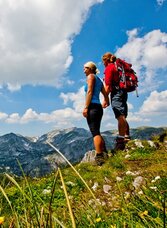 Zwei Wanderer mit Rucksäcken stehen auf einem Berggrat und blicken über eine alpine Landschaft mit Bergen, Wiesen und blauem Himmel
