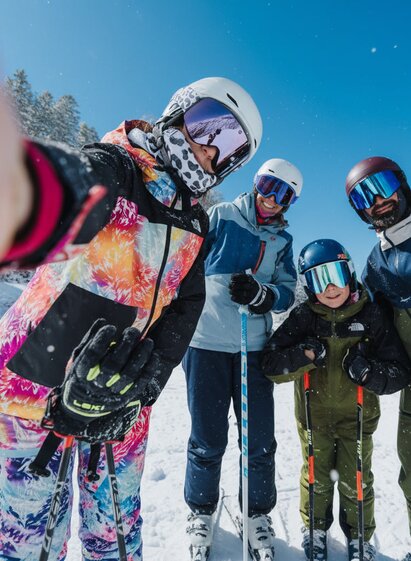 Eine Familie steht in Skibekleidung mit Skiern auf einer verschneiten Skipiste und blickt in die Kamera. Das Mädchen im Vordergrund hält die Kamera für ein Selfie. Alle tragen Skihelme und Skibrillen. Im Hintergrund sind verschneite Bäume und ein klarer blauer Himmel zu sehen.