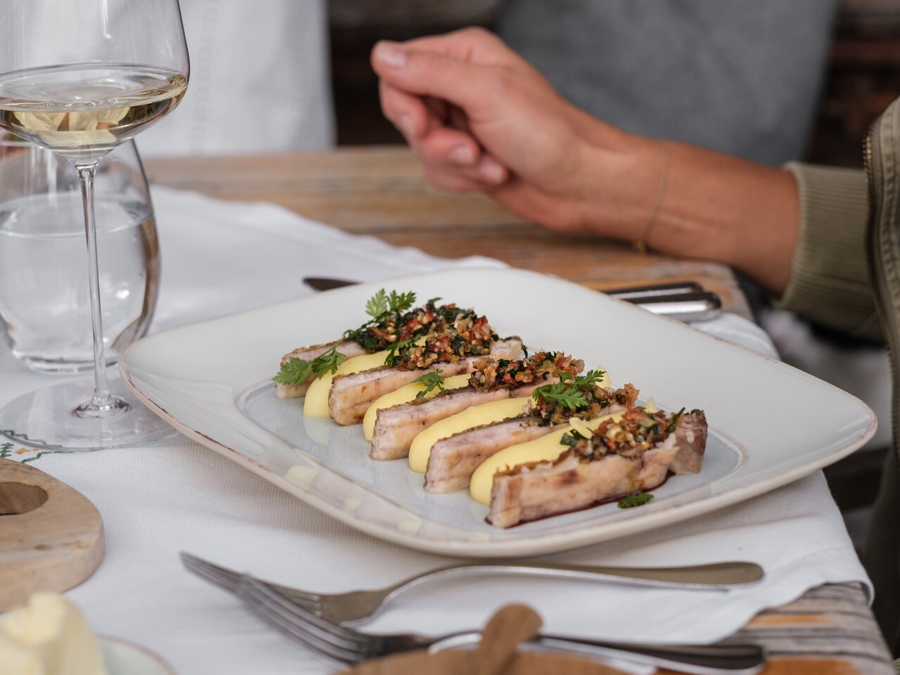 Several long pieces of a dish are arranged on a white plate, alongside yellow side dishes and a herb topping. Next to them, on a laid table, stand a glass of white wine and a glass of water.