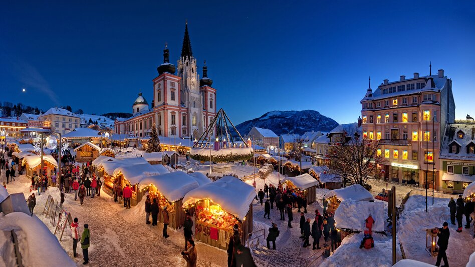 Der Hauptplatz Mariazell am Abend mit vielen Adventhütten und verschneiten Dächern, im Hintergrund die Basilika Mariazell und einigen Personen, die gemütlich durch die Hütten schlendern