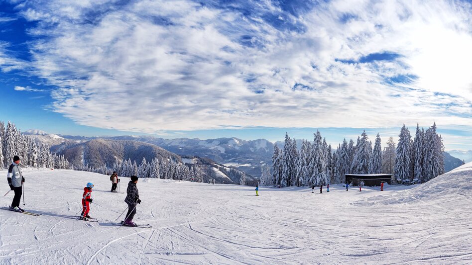 Ansicht einer Familie beim Skifahren auf der Mariazeller Bürgeralpe, im Hintergrund der Sessellift und verschneite Bäume