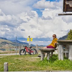 Radfahrerin in gelber Jacke sitzt vor einer urigen Berghütte auf einer Holzbank. Daneben steht ihr Mountainbike, im Hintergrund sind Berge, Almwiesen und ein bewölkter Himmel zu sehen.