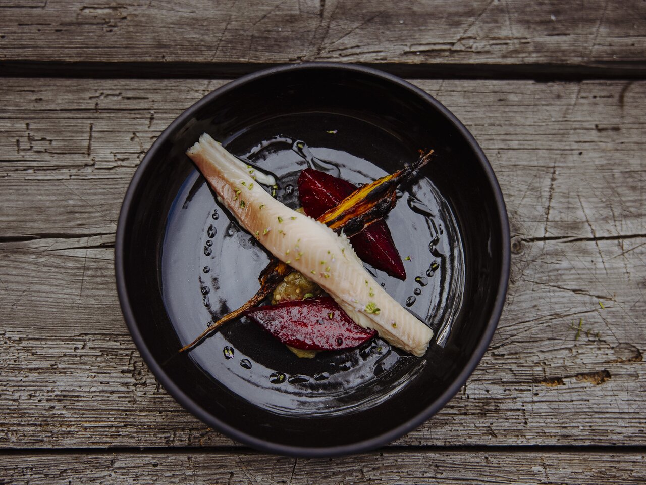 a fish dish, beautifully served on a black plate, with carrots and turnips, on a greyish wooden table