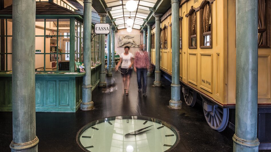 Eine Wartehalle im alten Stil, eine große Uhr ist im Boden integriert, große runde Leuchten hängen von der Decke. 