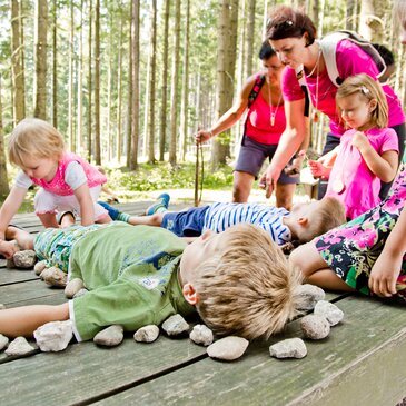 Zwei Kinder liegen auf einer Holzliege im Wald, Kinder legen Steine rundherum