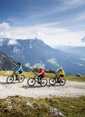 Three people ride their e-bikes up a gravel path to the alpine pasture, with a stunning mountain backdrop and scattered clouds in the background.