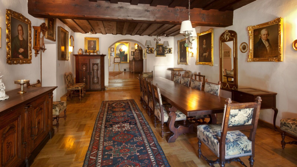 Eine prachtvoll eingericheteter Raum mit goldernen Bilderrahmen, eine langen Tafel mit gepolsteren Stühlen