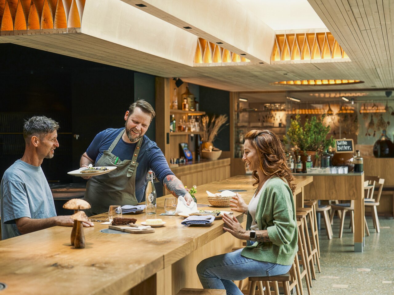 Ein freundlicher Kellner serviert einem Mann und einer Frau Essen an einer modernen Holztheke in einem stilvollen Restaurant mit warmem Licht und gemütlicher Atmosphäre.