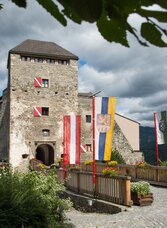 Ansicht vom Eingangsbereich der Burg Oberkapfenberg im Sommer, geschmückt mit Fahnen und mit schönem Blumenschmuck, im Hintergrund sieht man dunkle, stimmungsvolle Wolken am Himmel
