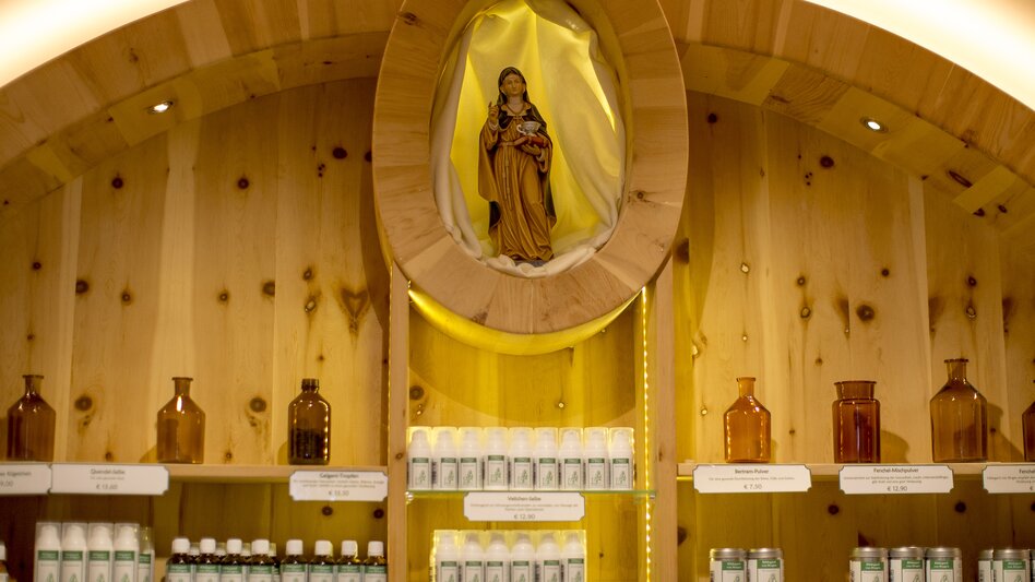 Ein hölzernes Regal mit verschiedenen Fläschchen und Dosen ist warm beleuchtet. In einer ovalen Nische oberhalb der Regale steht eine religiöse Figur, die eine Frau in einem langen Gewand darstellt. Die Produkte sind ordentlich auf mehreren Ebenen angeordnet, vor einer Wand aus hellem Holz.