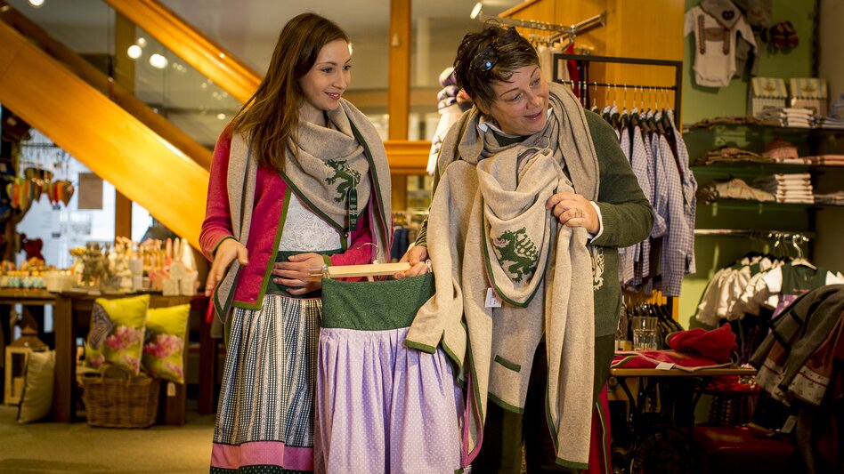 In einem gemütlich eingerichteten Trachtengeschäft steht eine junge Frau in traditioneller Tracht, bestehend aus einem Rock, rosaner Weste und einem grauen Schal. Eine weitere Dame steht beratend zur Seite und hält einen Rock in einer Hand und in der anderen eine graue Weste und einen grauen Schal, der mit dem steirischen Panther verziert ist. Der Laden ist warm beleuchtet, im Hintergrund sind weitere Trachten und Modeartikel auf Regalen und Kleiderstangen zu sehen. Eine Holztreppe führt zu einer oberen Etage.