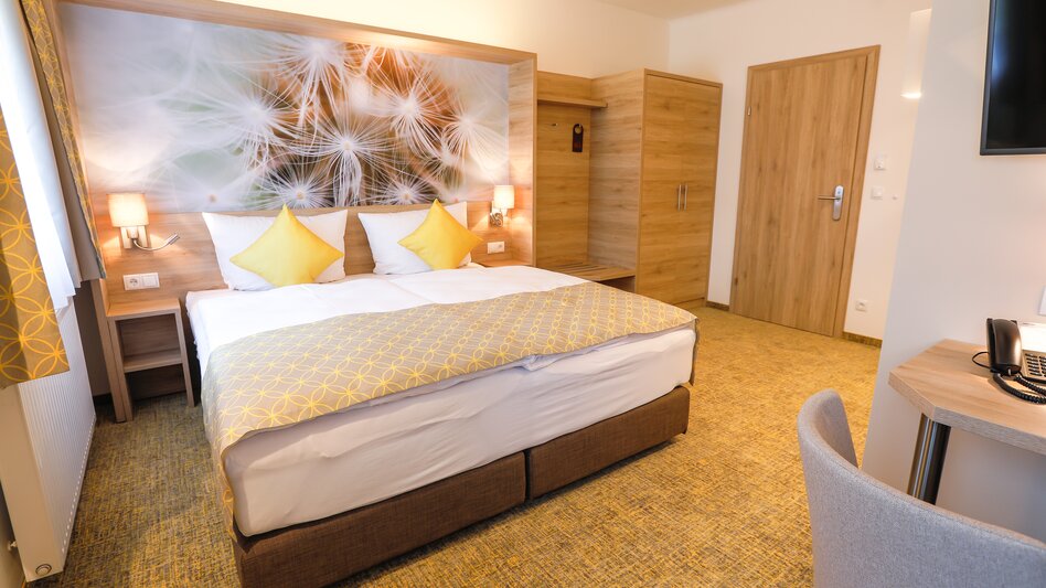 Ansicht eines Doppelzimmers im R&R Residenzen Hotel Mitterbach mit ocker-farbenem Teppichboden, einem Doppelbett, schönem, modernen Holzmobiliar und einem großen Bild an der Wand hinter dem Bett.