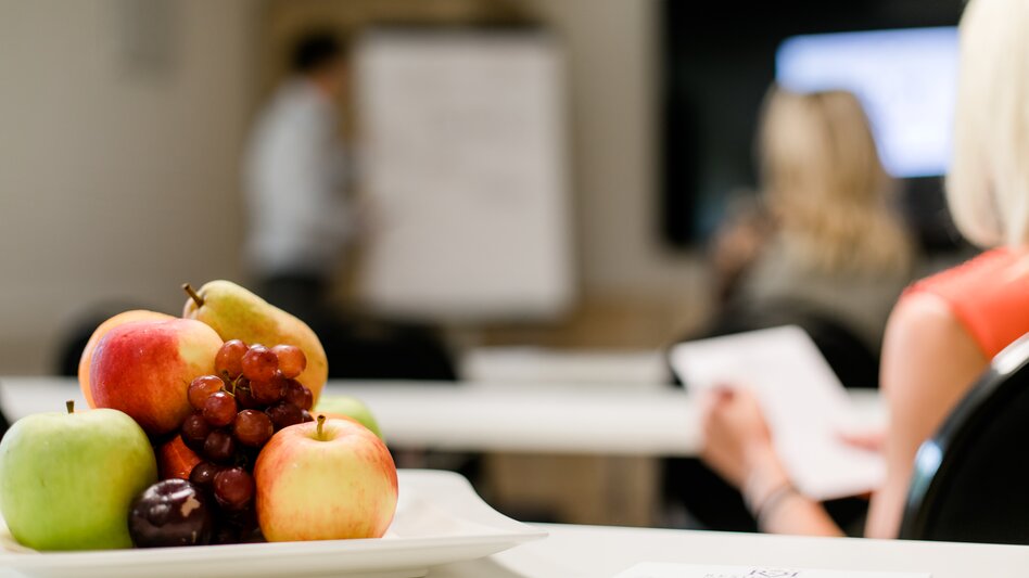 Nahaufnahme eines Obsttellers mit Äpfel, Birnen und Trauben, im Hintergrund sieht man Personen sitzen