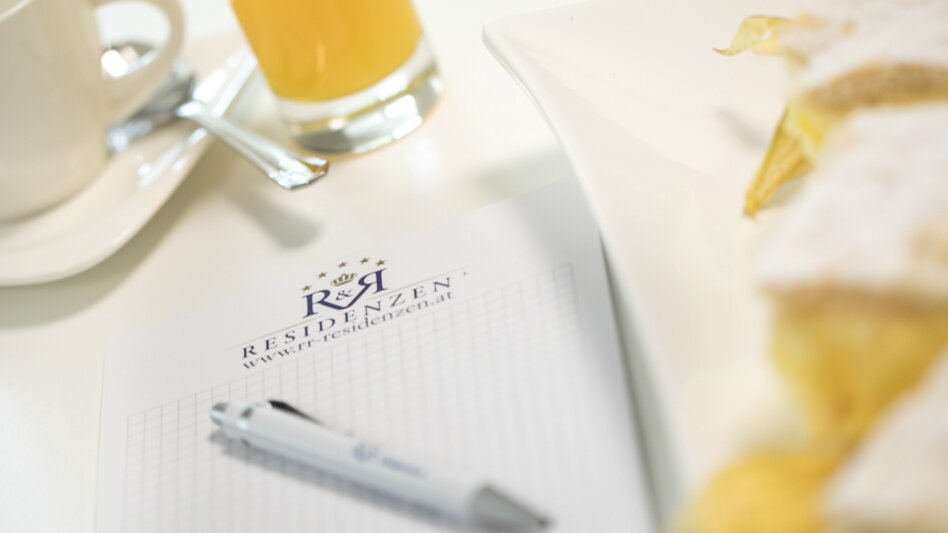 Auf einem Tisch liegen ein Notizblock und ein Kugelschreiben mit dem Logo des Hotels, davor steht ein Glas Orangensaft und eine Tasse Kaffee