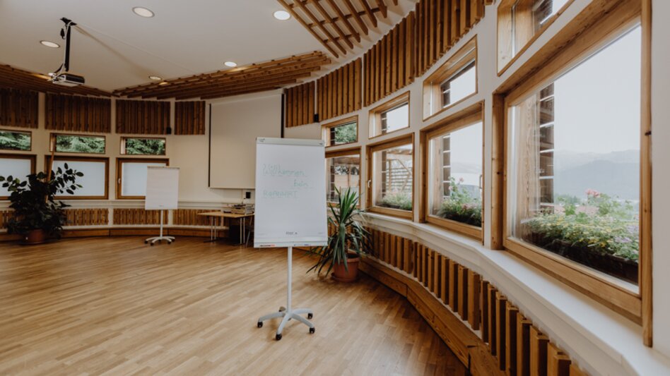Ansicht des Seminarraums mit vielen Holzelementen, grünen Pflanzen, Beamer, Leinwand und Flip-Charts.