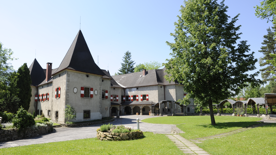Sommeransicht vom Schloss Pichl mit rot-weißen gestrichenen Fensterläden, umgeben von einer Wiese und Bäumen
