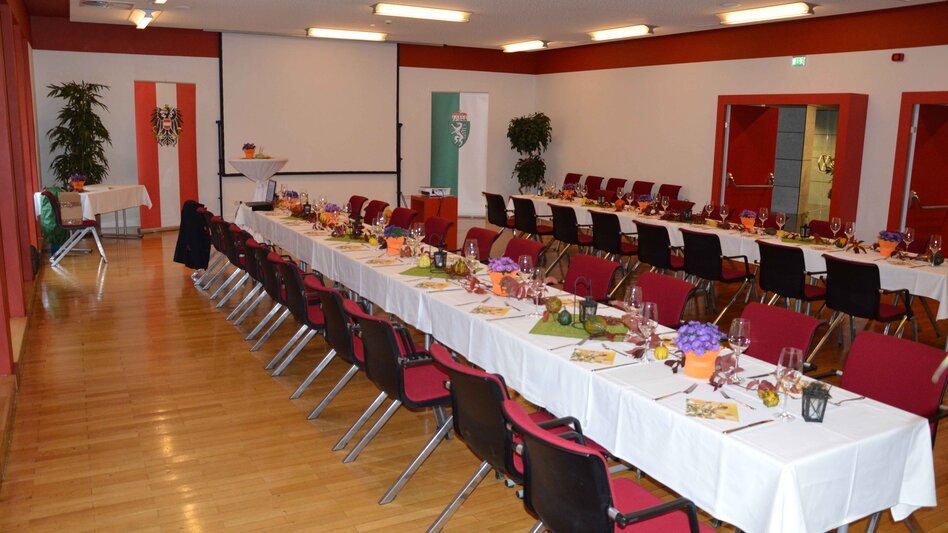 Ansicht des Kleinen Saals im Stadtsaal Mürzzuschlag mit 2 langen Tischreihen und roten Sesseln sowie einem Beamer mit Leinwand und Grünpflanzen als Dekoration