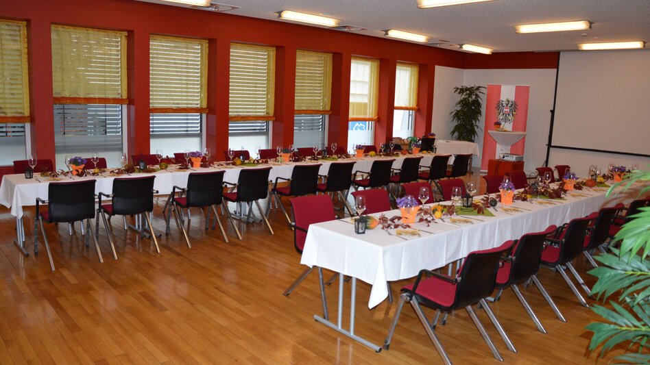 Ansicht des Kleinen Saals im Stadtsaal Mürzzuschlag mit mehreren Fenstern mit halb heruntergelassenen Jalousien, 2 langen Tischreihen und roten Sesseln sowie einem Beamer mit Leinwand