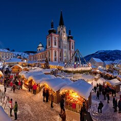 Winter Christmas market with beautifully lit huts in front of the basilica in Mariazell. Visitors stroll between the snow-covered stalls, while the church and snow-covered mountains can be seen in the background.