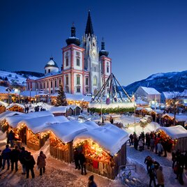 Christmas market in winter with atmospheric illuminated huts in front of the basilica in Mariazell. Visitors stroll between the snow-covered stalls, while the church and snow-covered mountains can be seen in the background.