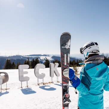 Eine Frau hält ihre beiden Skier in den Händen, im Hintergrund der Schriftzug des Skigebiets aus Eisen im Schnee