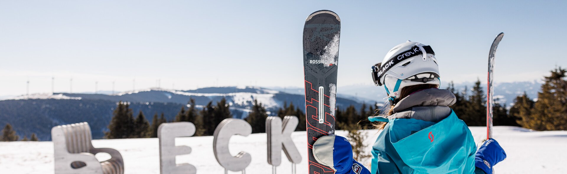 Eine Frau hält ihre beiden Skier in den Händen, im Hintergrund der Schriftzug des Skigebiets aus Eisen im Schnee