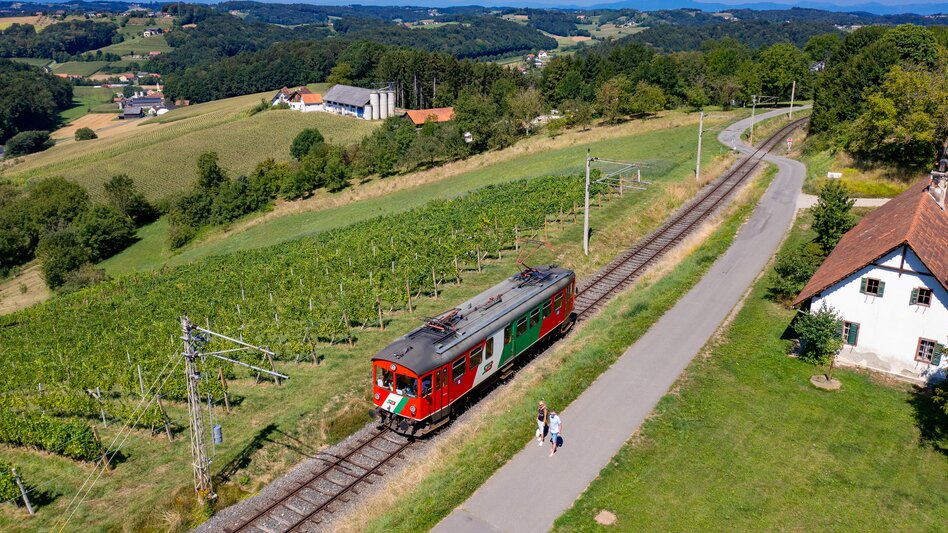 Die Gleichenbergerbahn ist aus der Vogelperspektive abgebildet, umgeben von der schönen Landschaft der Südoststeiermark. Links neben den Bahngleisen präsentieren Weinreben in frischem Grün. Im Hintergrund erblickt man die Wiesen, Felder und Wälder, bis die Bergkette auf den wolkenlosen Himmel trifft.  | © StB